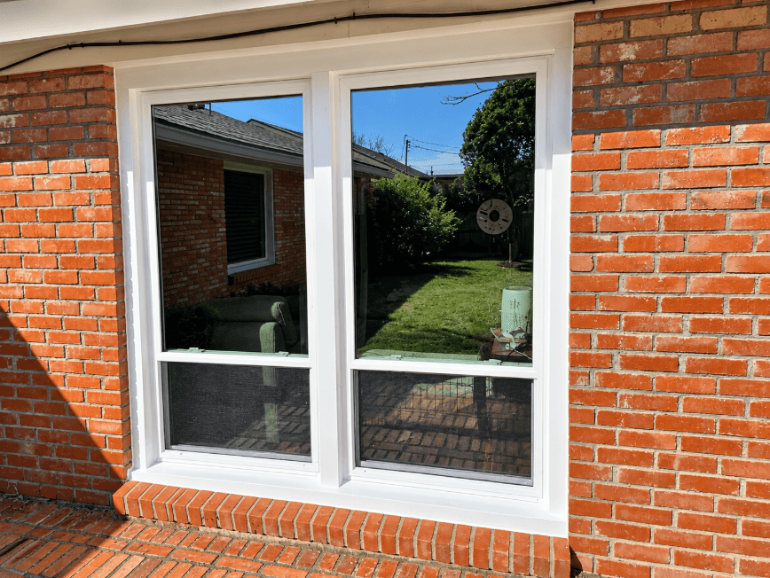 Twin white single-hung windows with a 70/30 sash split installed on a red brick home, overlooking a green backyard